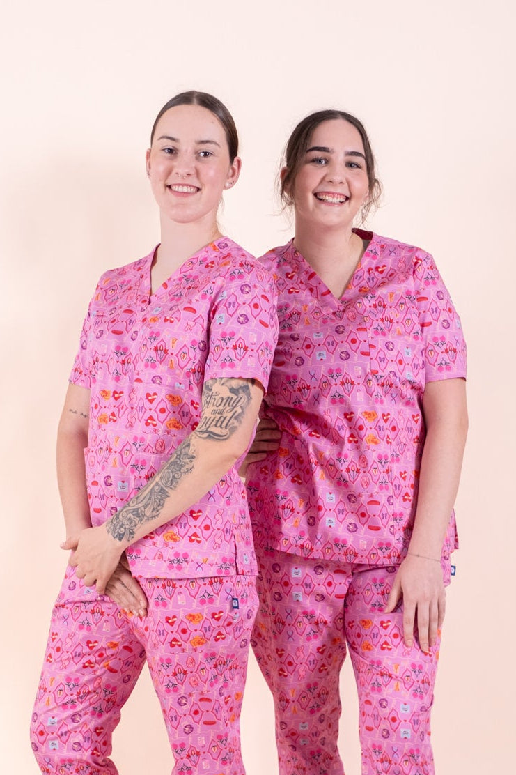 Two women modeling matching pink medical scrubs with a small floral pattern, standing against a solid pale peach studio background.
